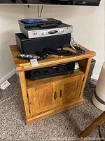 Front view of the wooden TV stand with electronics on top and open middle shelf containing equipment and a two-door cabinet below. Shows wood grain and wear.