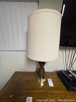 Front view of table lamp with turned wood base and tall drum fabric shade, visible damage on shade.