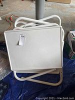 Folded tables stacked, showing white plastic tops and white tubular metal frames in folded state.