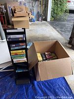 Photo showing a cardboard box filled with assorted VHS tapes alongside a tall black vertical VHS tape display stand.