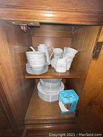 Image of upper wooden cabinet shelf showing stacked teacups, saucers, and small plates along with a box of wrapping material