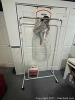 Metal clothing rack standing in a room against a white door. Some clothes hangers and a garment bag are on the rack. There is a box on the floor under the rack.