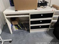Front and side view of white desk with 3 black drawers, keyboard pull-out shelf, and open compartment underneath. Desk surface has items on top and below showing usage and wear.