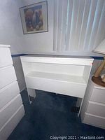 White twin size headboard showing front face with two open storage cubbies, placed in a carpeted room next to a white dresser and lamp.