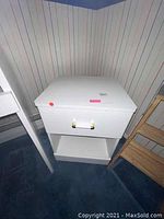 Photo showing the full white nightstand with drawer and open shelf located on a blue carpeted floor against striped wallpaper.