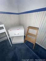White nightstand beside wooden step ladder against wall with wallpaper and blue carpet.