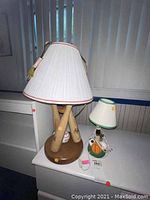 Photo showing two table lamps side by side; one with a base made of crossed wooden baseball bats and a white lampshade, the other smaller lamp with sports ball themed base including basketball and baseball with a white and green lampshade.