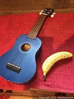 Blue ukulele laying on red fabric, seen in full front angle, nylon strings and white tuning pegs visible, with a banana included for size reference.