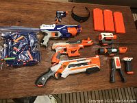 NERF collection including six blasters in white, orange, and blue, three orange magazines, black protective glasses, and darts scattered on a table.