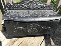 Front view of black ornate metal mailbox showing decorative scrollwork and floral embossed design, with chipped paint on top and sides.