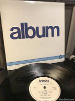 Front cover of the PIL 'album' vinyl record showing white background, blue text and record partially visible