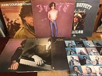 Photo showing seven classic rock vinyl records by artists including Van Morrison, John Cougar, Billy Joel, Joe Cocker, Ricky Skaggs, and Jimmy Buffett arranged on a surface.