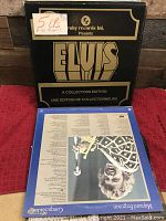Photo of the Elvis Presley 5-record box set with a black and gold collectors edition cover, alongside another record sleeve featuring an upside-down portrait on a blue background.
