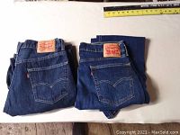 Two pairs of folded Levi Strauss jeans, both blue denim with leather brand patches on waistband, placed side by side with measuring tape above for scale.