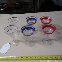 Front view of six cordial glasses, two each in red, white, and blue color bands, showing gold trim and clear stems, with a measuring tape for size reference.