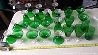 Top and angled view of large set of emerald green dessert and juice glasses with clear textured bases, arranged on white cloth.