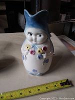 Front view of ceramic Toby jug with cheeky woman and flowers, showing approximate height with measuring tape.