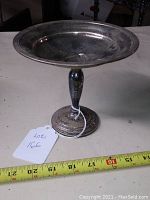 Front view of a vintage silver candy dish showing the embossed base and wide shallow bowl, placed on a surface next to a measuring tape showing approximately 8 inches in height.