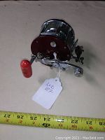 High angle view showing the chrome and maroon finish, the red handle knob, attached label, and the spool of the reel.