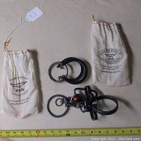 Two metal brain teasers displayed side by side with their branded cloth storage bags. A measuring tape shows size context.