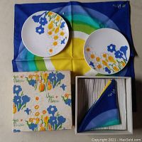 Two round plates with blue, yellow, and orange floral pattern, laid on one of four matching blue, green, yellow, and white concentric circle cloth napkins; original box with floral artwork and Vera Mikasa name in view