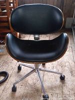 Front view of black padded seat and backrest with wooden shell outline and chrome caster base on wooden floor