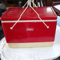 Front and side view of vintage red Coleman cooler with cream-colored base, white metal handle, and Coleman logo on front.