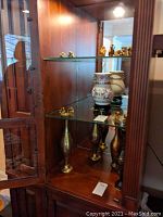 Wide shot of wooden glass display cabinet containing small Asian figurines, lattice vase with fish decoration, and multiple metal vases of differing heights.
