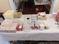 Overview of set including mugs, glass bowls, serving dish, porcelain demitasse set on tray, and other glassware on white tablecloth.