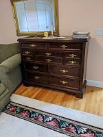 Full front angled view of the Bassett dresser with a dark wood finish, ornate gold colored drawer pulls, against a pale pink wall with a large gold framed mirror above.