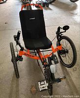 Front view of orange trike showing frame, mesh seat, clipless pedals and front wheels