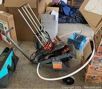 Photo showing Champion Wheelybird auto-feed skeet shooter trap with support arm, wheel, and power cord, surrounded by cardboard boxes and bags.