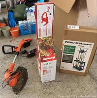 Whole lot showing Homelite hedge trimmer in box, Black & Decker weed wacker used, and Tiller & Rowe garden tool organizer boxed open and next to other items.
