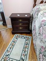 View of one dark wood nightstand with three drawers and brass handles, placed next to floral bedspread and small floral rug on hardwood floor.
