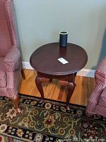 Wooden side table with round top, dark finish, cabriole legs, and a small drawer, sitting between two upholstered chairs on a patterned rug.