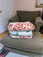 Three folded wool blankets stacked on a green upholstered chair. The top and bottom blankets have a prominent orange swirl pattern, with a cream base. The middle blanket appears white with green leafy or branch-like patterns.