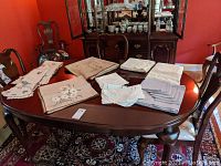 Overview photo of all table linens on a wooden dining table, showing folded fabric pieces including embroidered and hemstitched linens in mostly light neutral tones.
