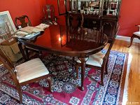 Photo showing the dark wood high gloss oval dining table with two chairs around it and red patterned carpet underneath. Reflection of chandelier visible on tabletop.
