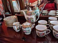 11 cups, 11 saucers, teapot, sugar bowl, and creamer arranged on wooden table, showing decorative floral and oriental patterns with gold trim