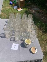 Photo showing rows of clear glass mason jars arranged on table outside in sunlight. 17 large jars visible, with 1 small and 2 extra small jars in front. One small jar with metal lid is present with another lid beside it.