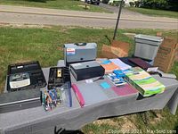 Table with various office supplies laid out including label printer, cashbox, files, pens, and stationery items