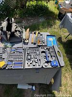 Wide view of table showing socket sets, hammers, grease gun, sharpening kit, and other hand tools.