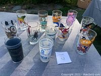 Full set of 11 assorted collectible juice glasses including McDonald's and location/military themes on outdoor table.