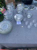 Overview of the entire lot arranged on a gray cloth surface outdoors. Shows stacks of clear bowls, dessert plates, salad plates, juice glasses, one carafe, and one bowl.