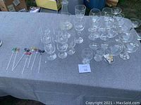 Photo of assorted clear barware glasses including champagne flutes, wine glasses, monogrammed drink glasses, Baileys glasses, and drink stirrers on a gray tablecloth outdoors.
