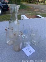 Full lot of one large milk carafe, two small milk carafes, and five glasses arranged on table outdoors