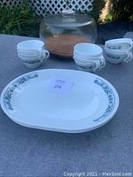 Display of 4 oval Corelle dishes with blue floral pattern, 14 matching Corelle mugs, covered glass platter with wooden base on table outdoors.
