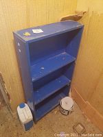 Blue wooden bookshelf placed against a damaged wall with peeling wallpaper, shows three shelves, surface wear and dirt, with paint chipping.