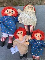 Photo showing five Raggedy Ann dolls and one Raggedy Andy doll outdoors, laid on a flat surface. Dolls have red yarn hair, painted or stitched facial features, dressed in various fabrics including floral and pastel dresses with striped stockings.