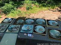 11 decorative plates arranged on a table outdoors showing maritime scenes and gold rims